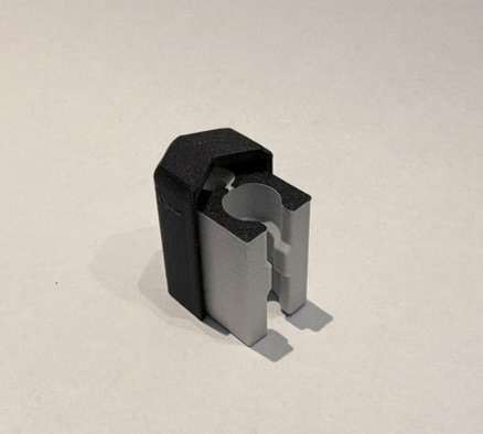 1-weg eindconnector: lichtgrijs 3D-geprint lichaam met halfronde groef naar de rail, zwarte buitenkap die het kanaal aan één kant afsluit, gezien in driekwart van bovenaf op een lichte achtergrond